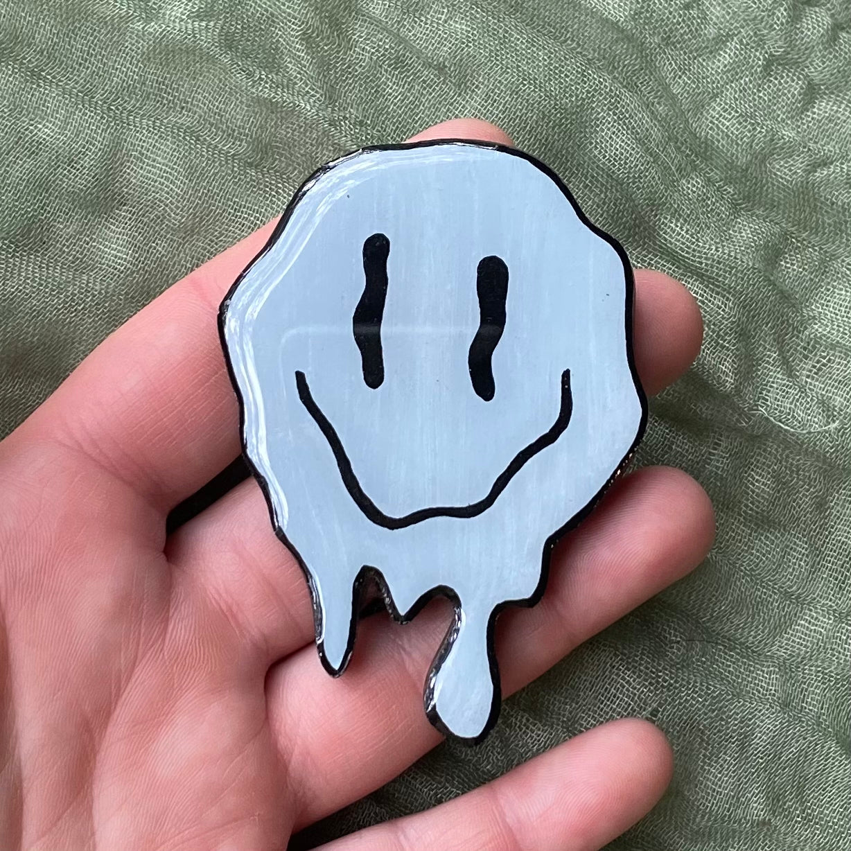 This magnet that is shaped like a melting smiley face. The top is round and the bottom has drips coming down as if it is melting. The magnet is painted light blue and the eyes and smile are painted to look like they are melting as well. 