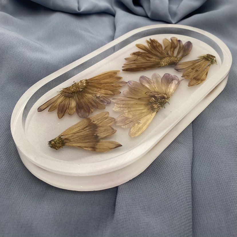 Photo shows an oval-shaped resin tray with several segments of withering daisies in front of a white background.