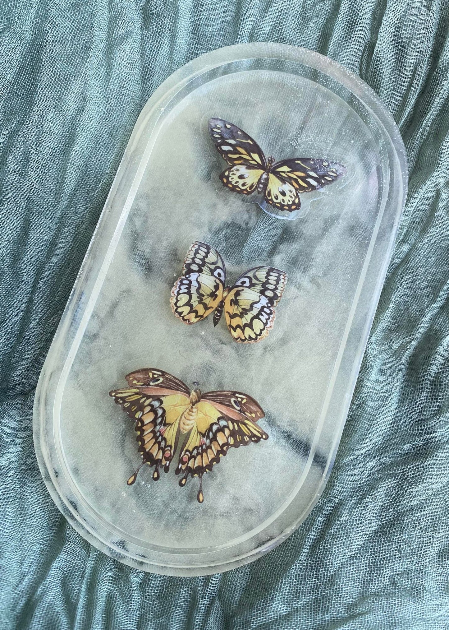 Photo shows an oval-shaped resin tray with three yellow butterfly stickers inside in front of a smoky yellow background.