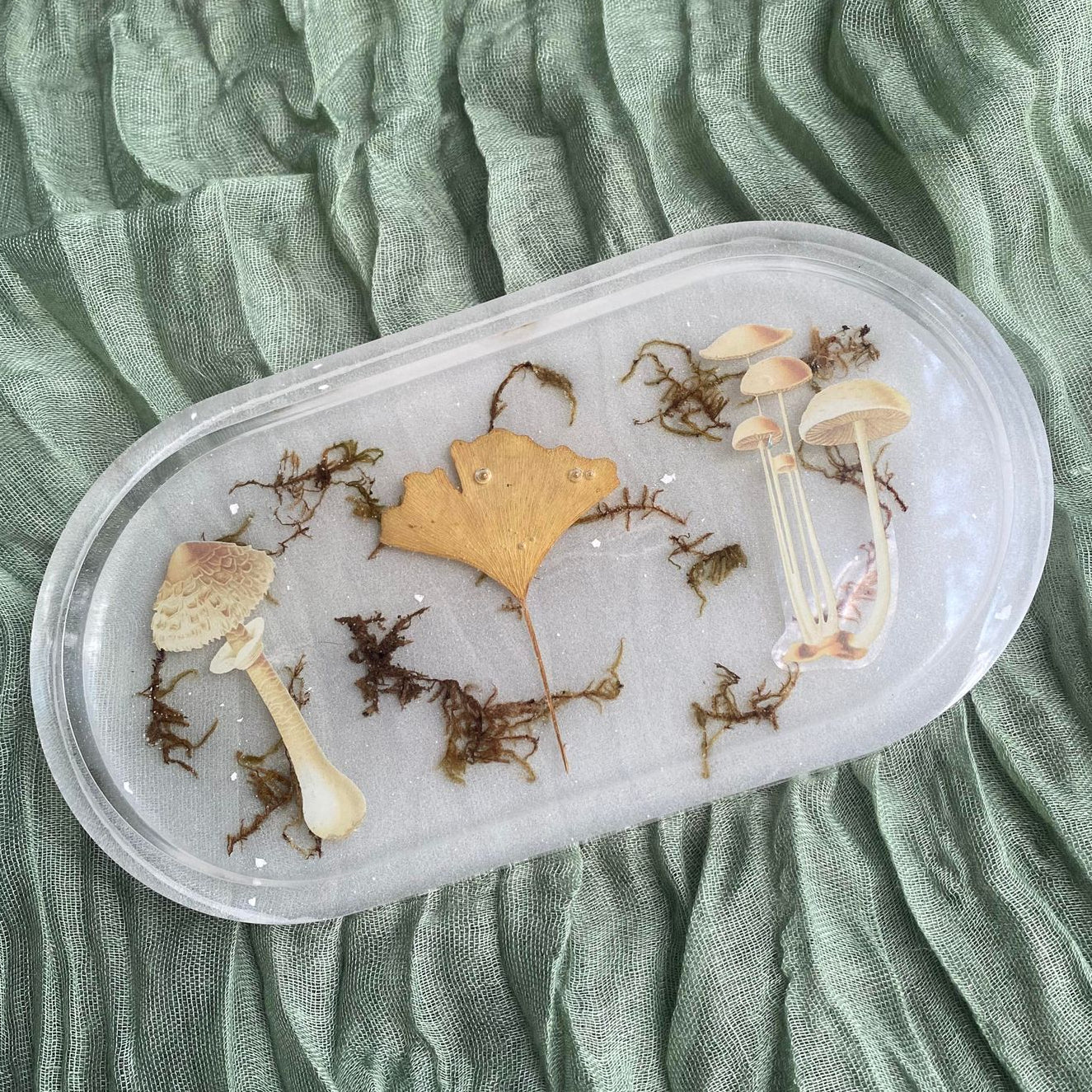 Photo shows an oval-shaped resin tray with a ginkgo leaf, mushroom stickers, and moss inside in front of a white background.