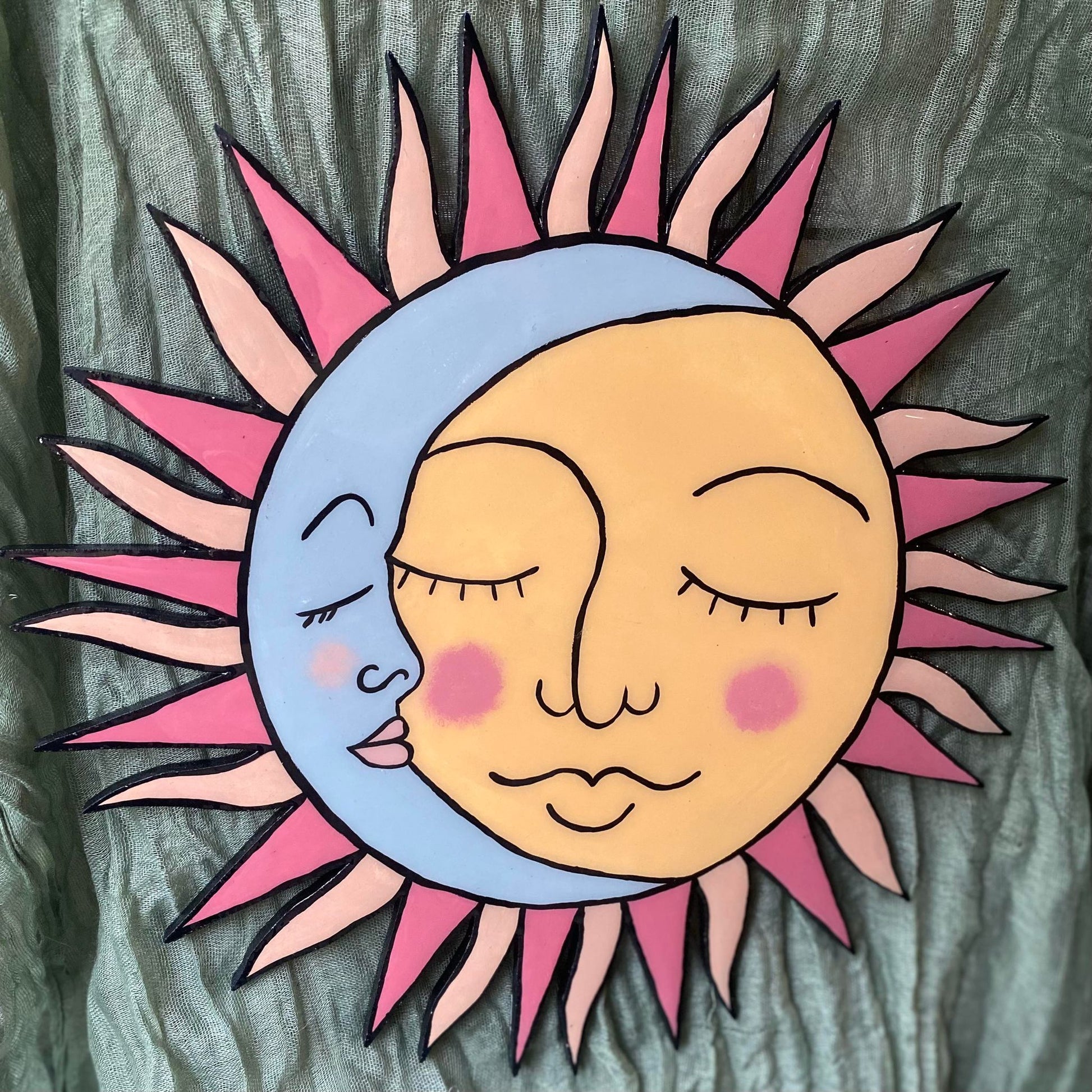 The wall hanging is a yellow sun with alternating pointed and wavy rays coming from the center. The rays also alternate between a shade of light orange and light red. On the left side of the sun, overlapping the face a bit is a blue crescent moon. It is seen from the side, so the right side of the moon's face is all that is visible. Both the sun and moon look serene and have their eyes closed as if they're asleep.