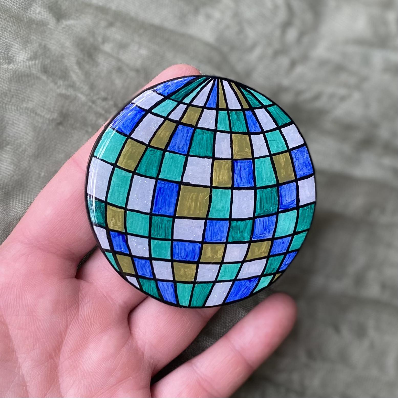 This disco ball magnet is round with black outlines. Each mirror is painted a different shade of blue or green, and the lightest blue mirrors also have a layer of glitter paint. 