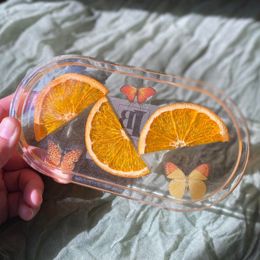 Photo shows a hand holding an oval-shaped clear resin tray with real orange segments and three orange butterfly and moth stickers inside.