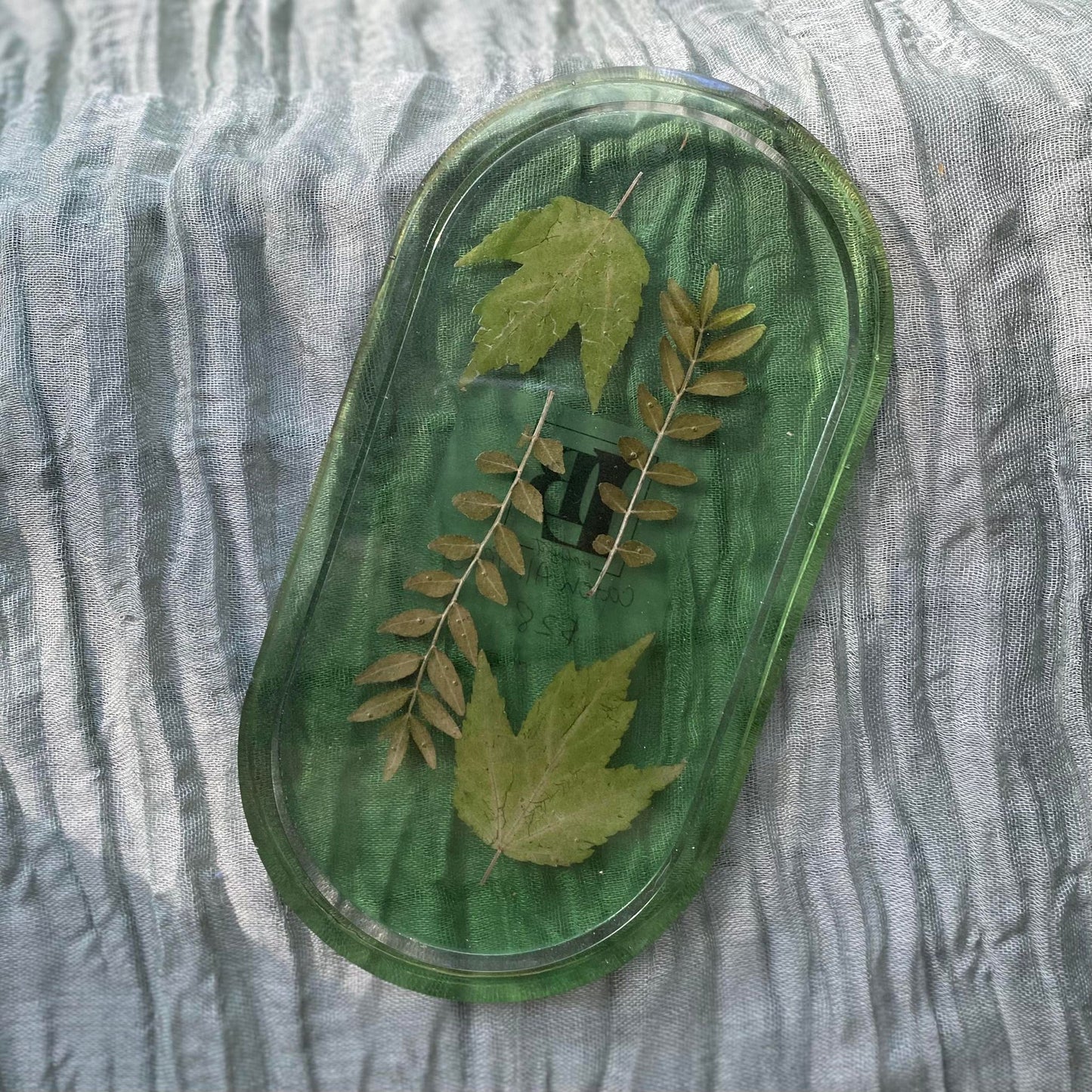 Photo shows an oval-shaped resin tray with dried greenery inside in front of a transparent green background.