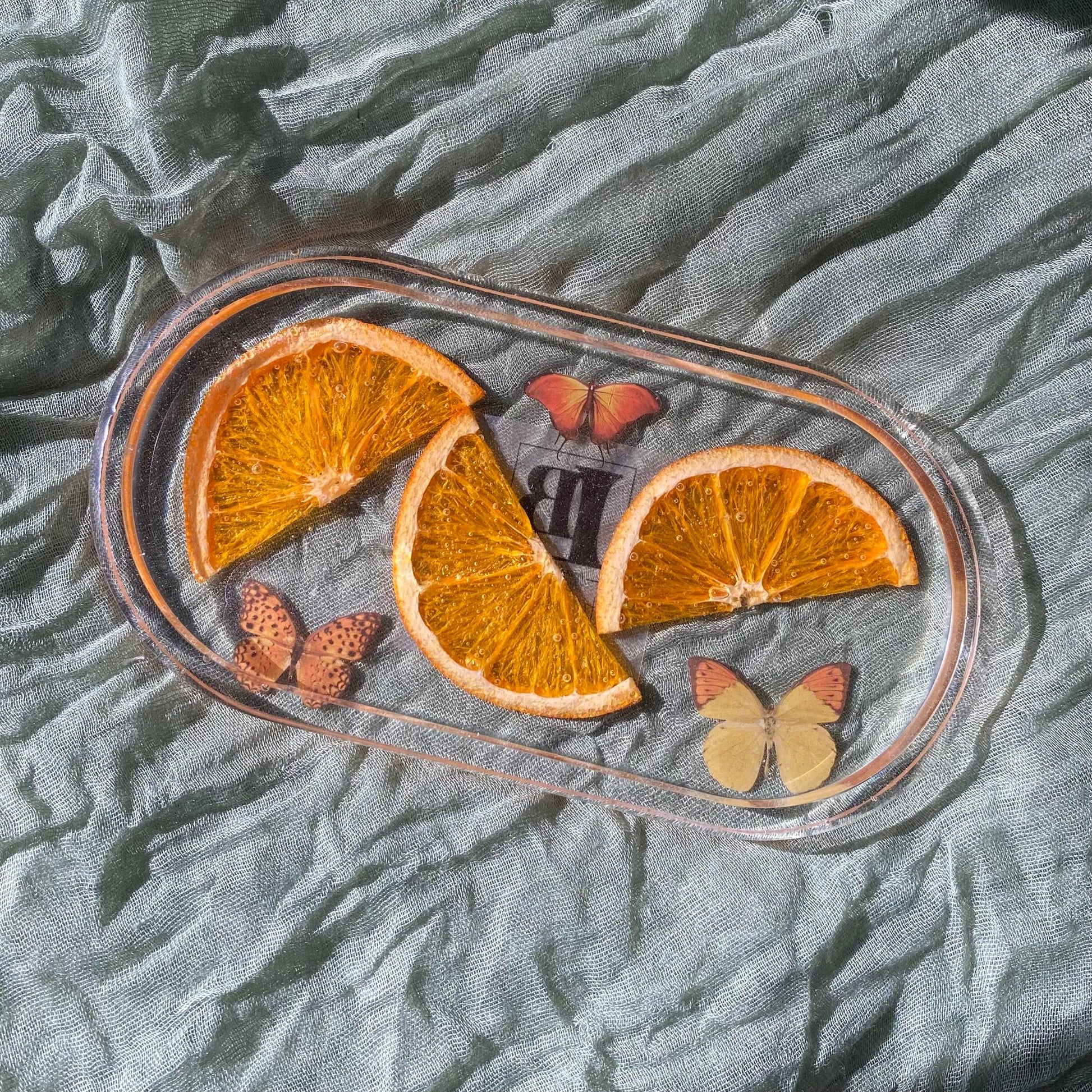 Photo shows an oval-shaped clear resin tray with real orange segments and three orange butterfly and moth stickers inside.