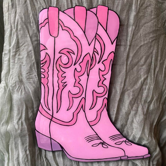 This wall hanging is cut in the shape of a pair of cowboy boots, with the boot on the left in the foreground and the boot on the right slightly hidden in the background. They are painted in shades of pink with black outlines, in the style of American traditional tattoos. They have a classic stitching design on the top half, a medium sized heel, and pointed toes.