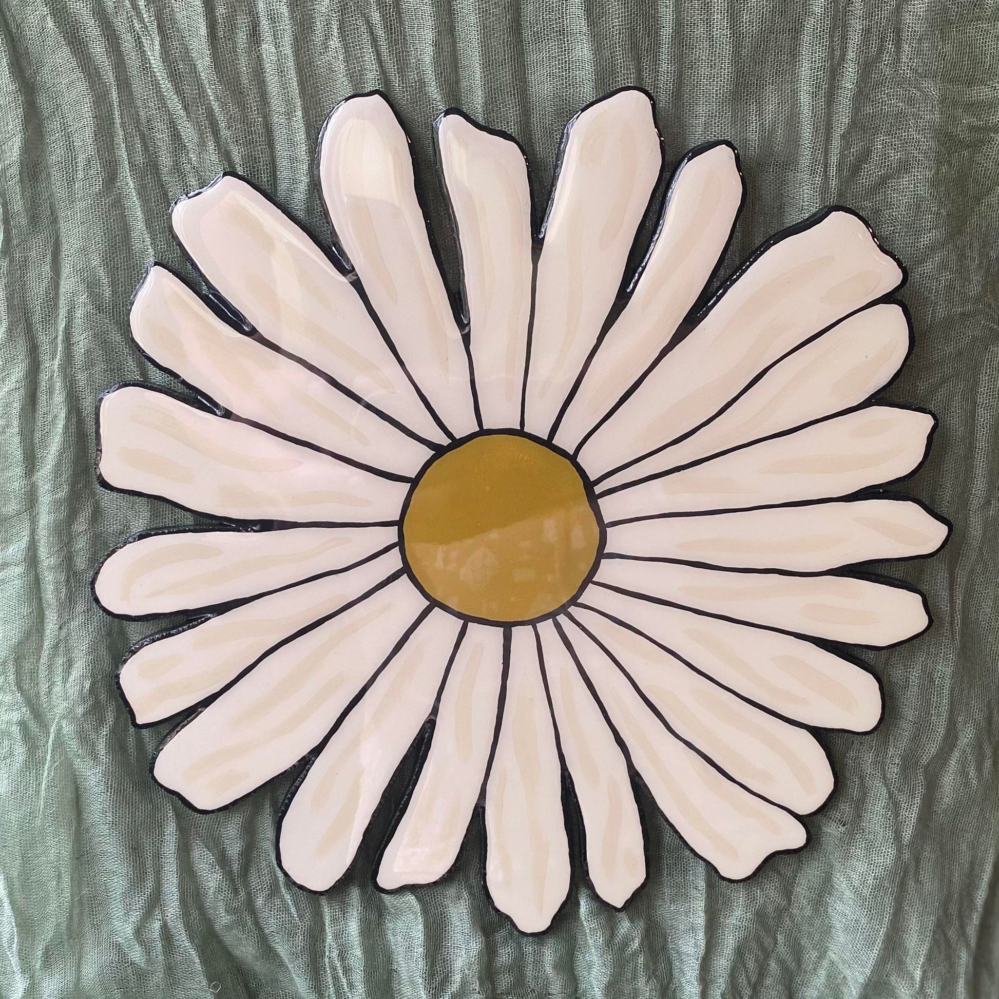 This wall hanging is in the shape of a daisy. The petals are white with ivory highlights to add depth, and the center of the flower is a mustard yellow. The daisy is painted in the style of American traditional tattoos, so it has bold black outlines.