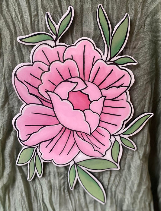 This wall hanging is in the shape of a peony flower drawn in the style of American traditional tattoos. There are a few leaves sticking out from behind the flower petals, which are painted in a few shades of light olive green. The peony is painted in shades of pink with black outlines.