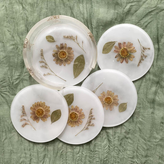 Top view of all the coasters and the matching holder. Each coaster is round with a white background, a pale purple flower in the center, and a piece of baby's breath and a leaf inside. The holder is similar but it features extra leaves and baby's breath.