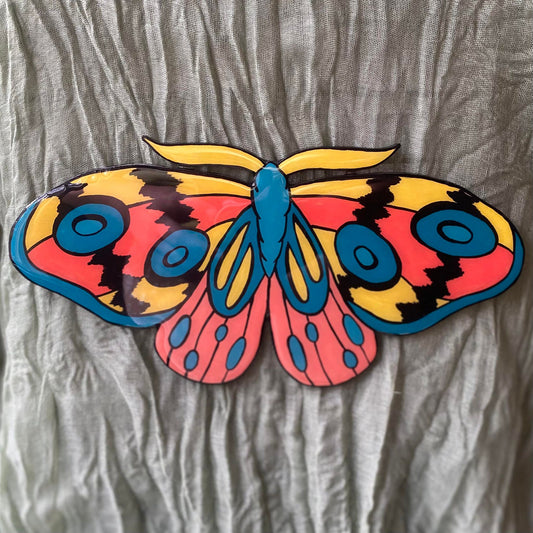 This wall hanging is cut in the shape of a moth. It is painted with yellow, teal, and coral. The antennae are yellow, the body is teal, and the wings have detailed designs on them. The top and bottom wings both have abstract designs on them, using coral, teal, and yellow. The moth is symmetrical from left to right, and everything is outlined in black.