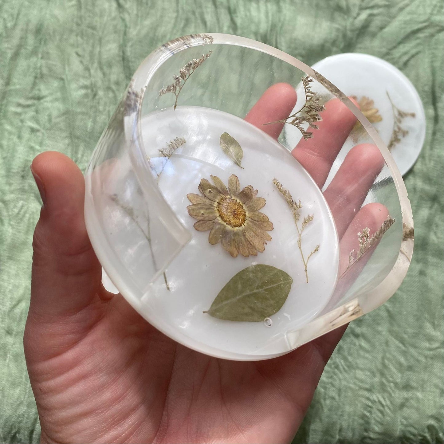 Close up of a round coaster holder which has a white background, a pale purple daisy in the center and various greenery surrounding it. The sides of the coaster holder are clear and have pieces of baby's breath inside.