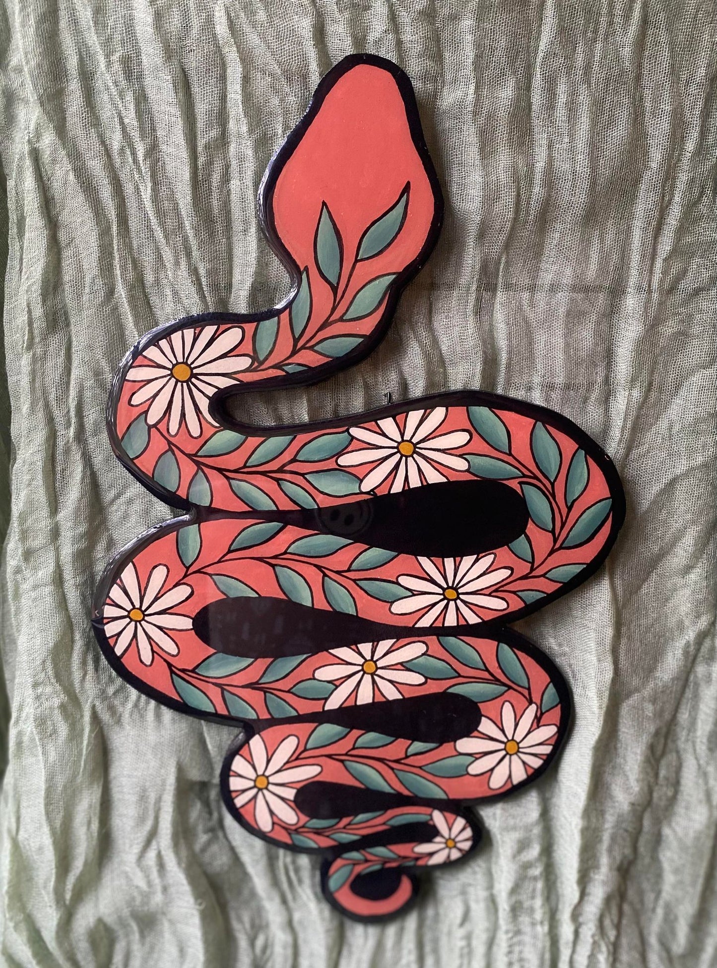 This wall hanging is cut out in the shape of a slithering snake. It is painted rust orange with white and yellow daisies and vines of green leaves trailing over it. 